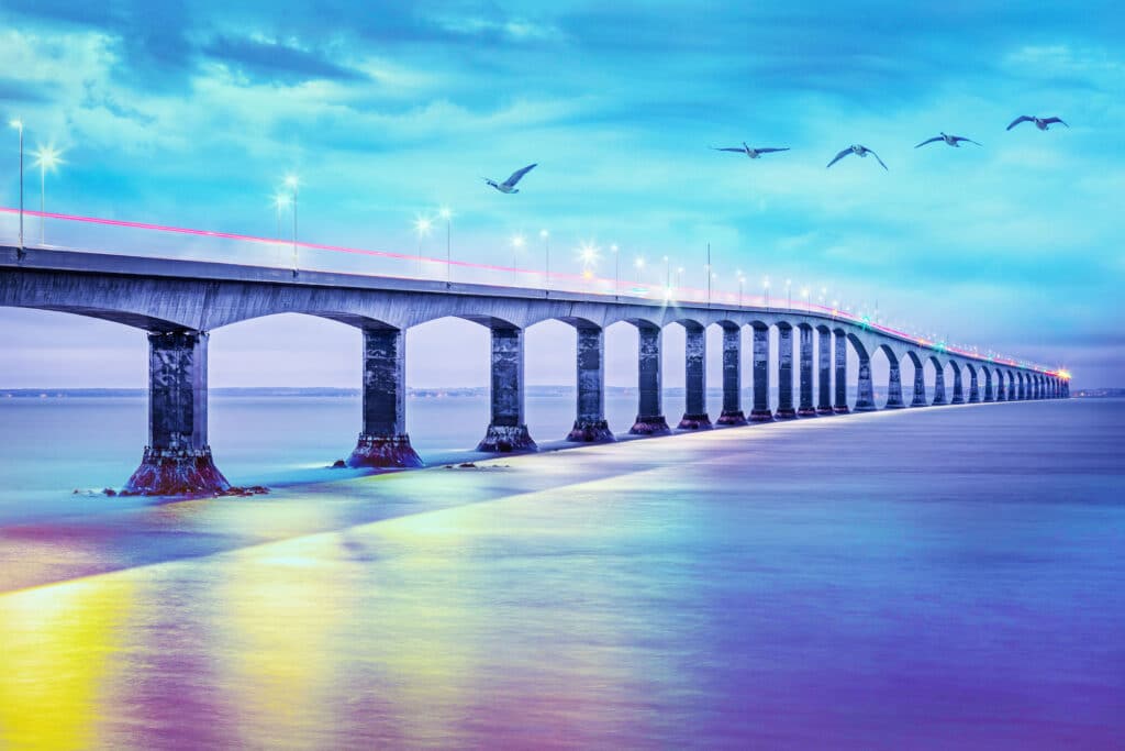A long bridge spans over calm water under a vibrant, colorful sky. Several birds fly above the bridge.