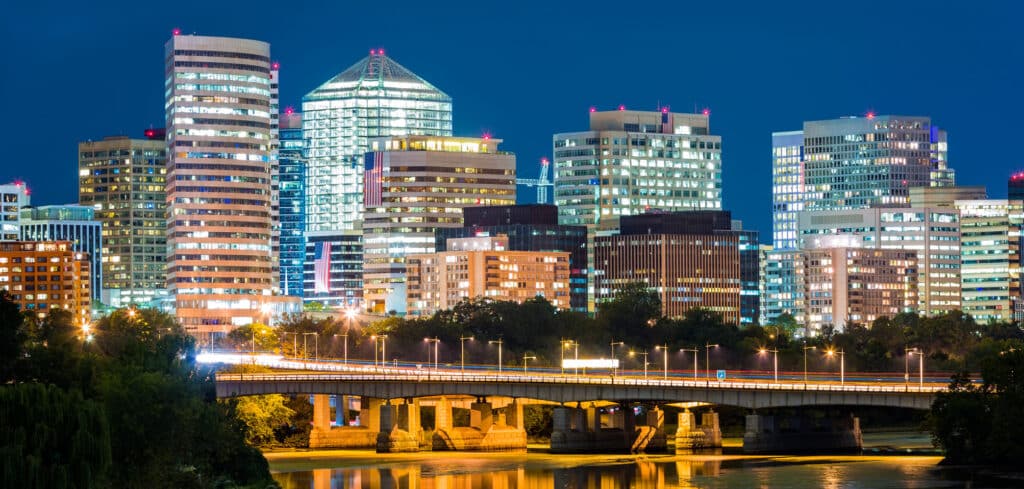 Night view of a city skyline with illuminated high-rise buildings and a bridge over a river in the foreground.