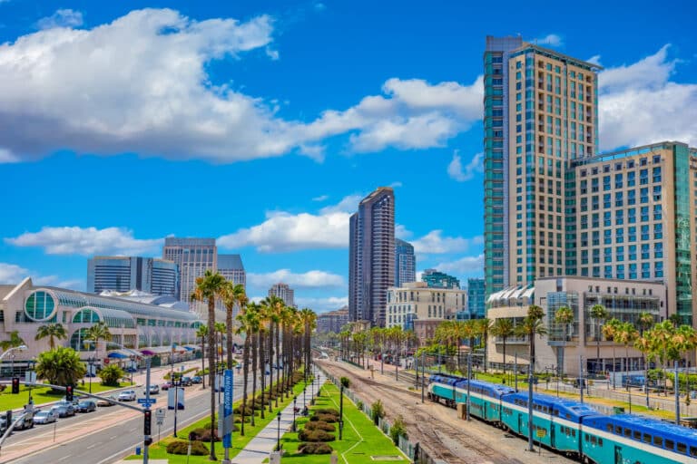 Cityscape featuring tall buildings, a train on tracks, palm-lined streets, and a clear blue sky with clouds.