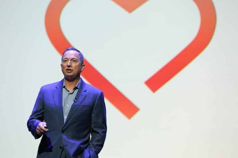 A man in a suit gives a presentation in front of a graphic resembling a heart on a screen.