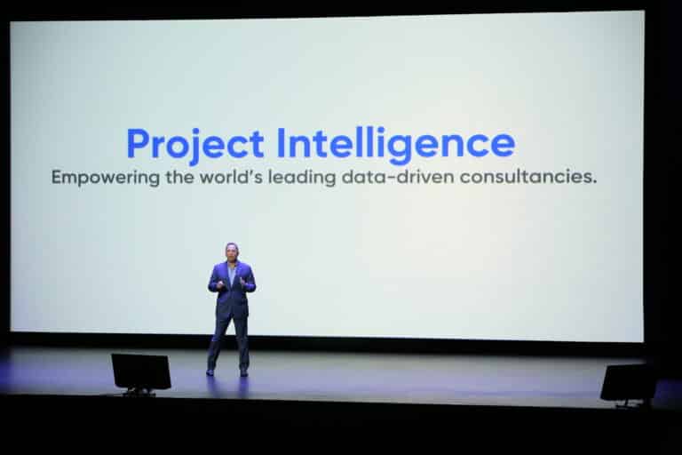 A person stands on a stage in front of a large screen displaying "Project Intelligence: Empowering the world's leading data-driven consultancies.