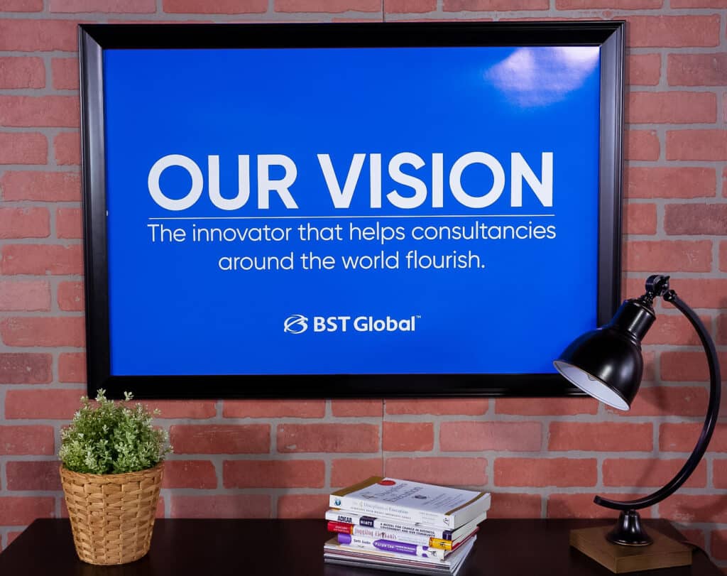 A framed sign on a brick wall reads, "OUR VISION: The innovator that helps consultancies around the world flourish," with "BST Global" below. A lamp, books, and a potted plant are on a table nearby.