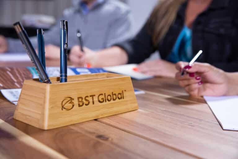 People working at a table with notepads and pens; a wooden pen holder with "BST Global" engraved.
