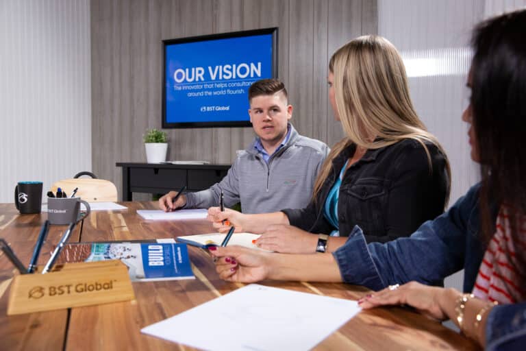 Three people sit at a conference table discussing in a meeting room with a screen displaying "Our Vision" in the background.