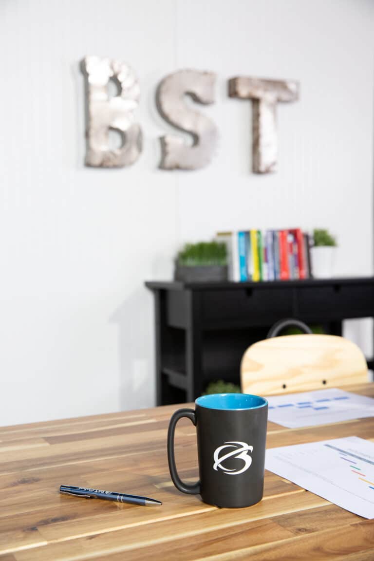 A wooden table with a pen, a mug, and a paper. Books and potted plants are on a black shelf in the background. Silver letters "BST" are mounted on the wall.