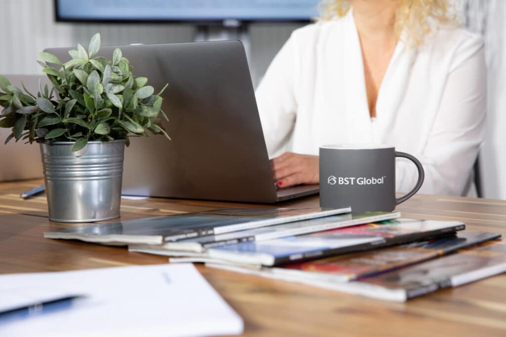 A person works on a laptop at a table with a potted plant, a "BST Global" mug, and a stack of magazines.
