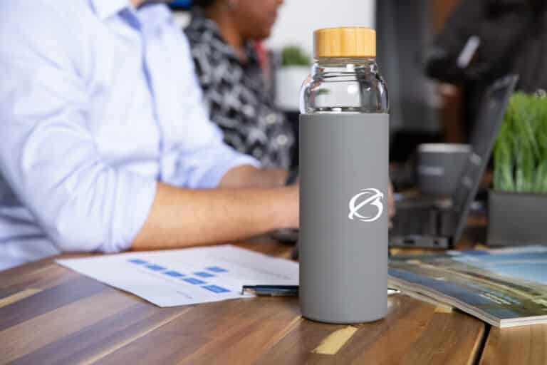 A glass water bottle with a gray sleeve sits on a wooden table near a laptop and papers.