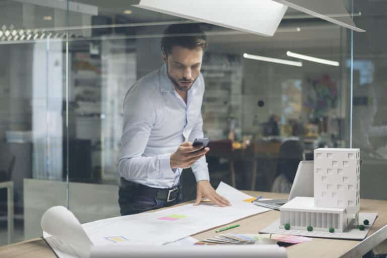 A man in a white shirt stands in an office, looking at his phone. He is surrounded by architectural plans and a model building on the desk.