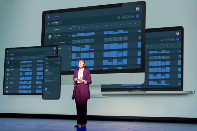 A person stands on stage in front of a large screen displaying a digital dashboard on various devices, including a laptop, tablet, and smartphone.