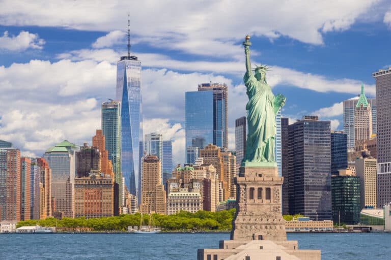 The Statue of Liberty stands in the foreground with the New York City skyline, including One World Trade Center, in the background on a clear day.