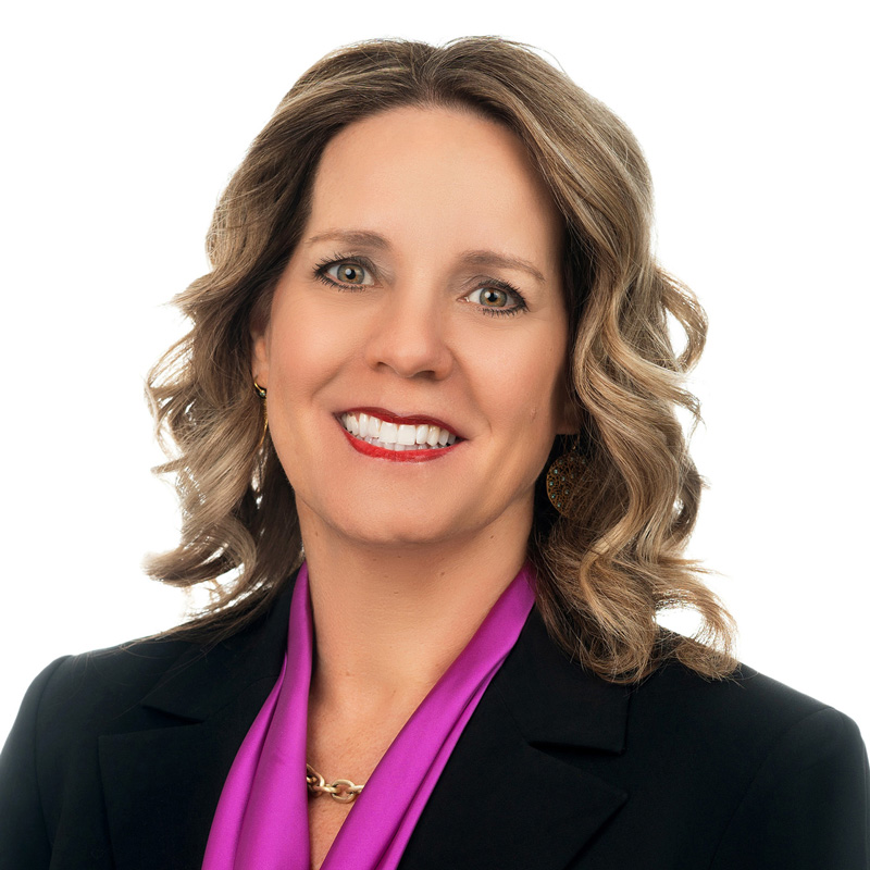 Eileen M. Canady with shoulder-length wavy hair, wearing a black blazer and a purple scarf, smiling at the camera against a white background.