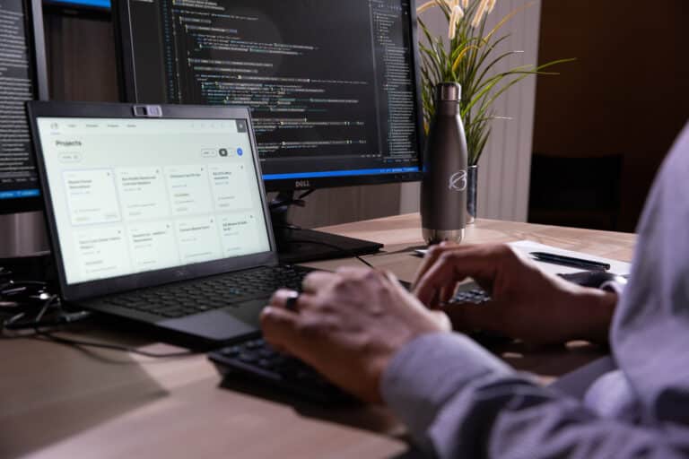 A person types on a keyboard at a desk with two monitors. One screen shows code; the other displays a project management tool. A water bottle and plant are nearby.