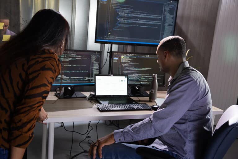 Two people working in an office, examining multiple computer screens filled with code and data.