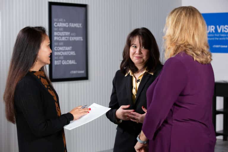 Three women stand and talk in an office setting. One holds a clipboard. A framed sign is visible in the background.