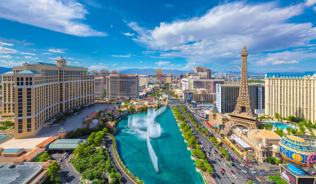Aerial view of Las Vegas Strip featuring hotels, casinos, and the Bellagio fountain with cityscape and Ferris wheel in the background under a blue sky.