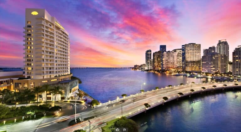 A vibrant cityscape at sunset, featuring skyscrapers along a waterfront with a lit bridge connecting two sides. The sky is filled with colorful clouds, and the water reflects the vibrant hues.