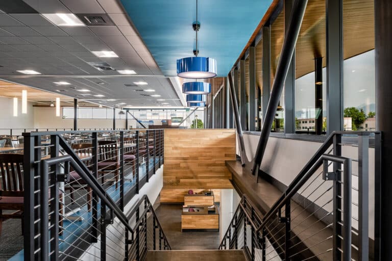 A modern interior with metal railings, wooden accents, blue ceiling, and large windows. The space includes an upper area with tables and a staircase leading to a lower level.
