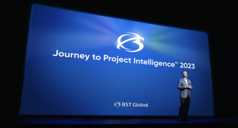 A man stands on stage next to a large screen displaying "Journey to Project Intelligence™ 2023" with the BST Global logo.