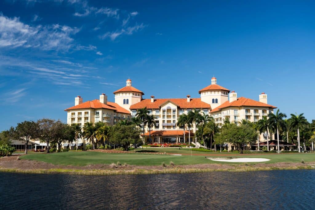 A large, luxurious building with orange roofs is surrounded by palm trees near a body of water under a clear blue sky.