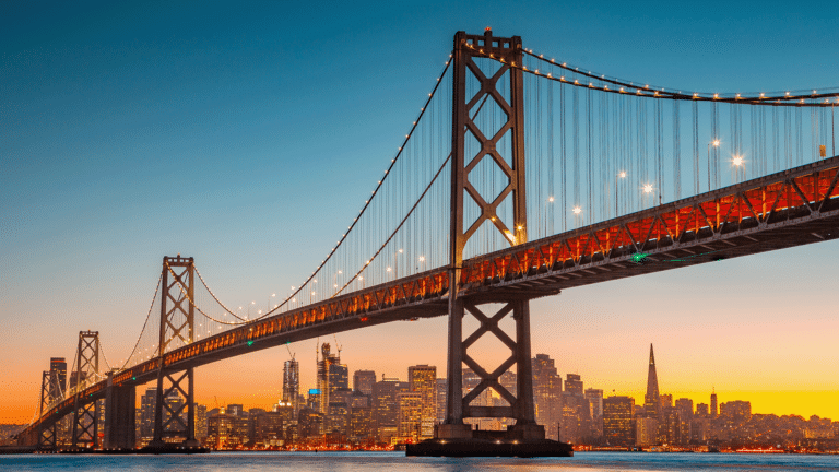 A suspension bridge with illuminated cables spans over water at sunset, with a city skyline in the background.