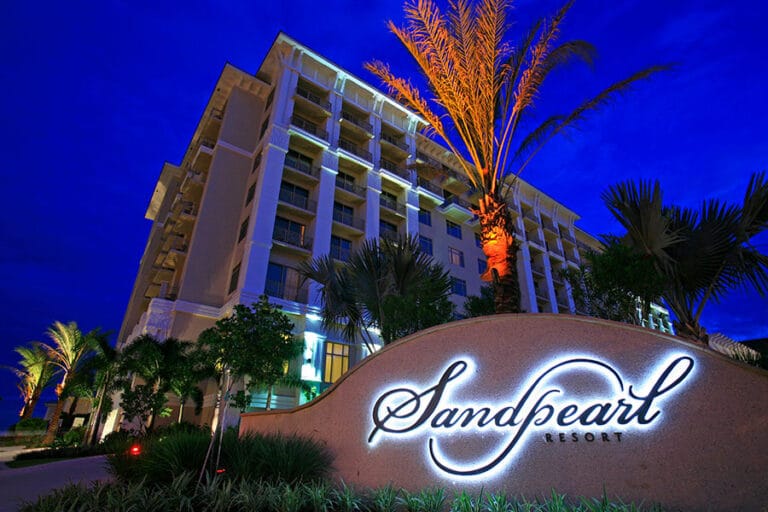Night view of a multi-story hotel with palm trees in front, featuring a lit "Sandpearl Resort" sign with a blue sky backdrop.