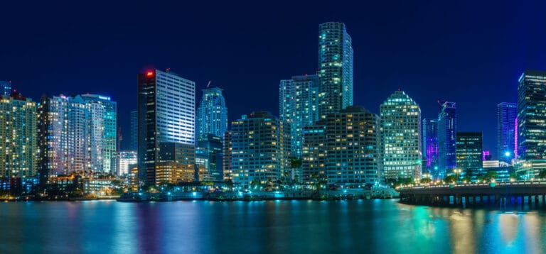 City skyline at night with illuminated skyscrapers reflecting on the water below.