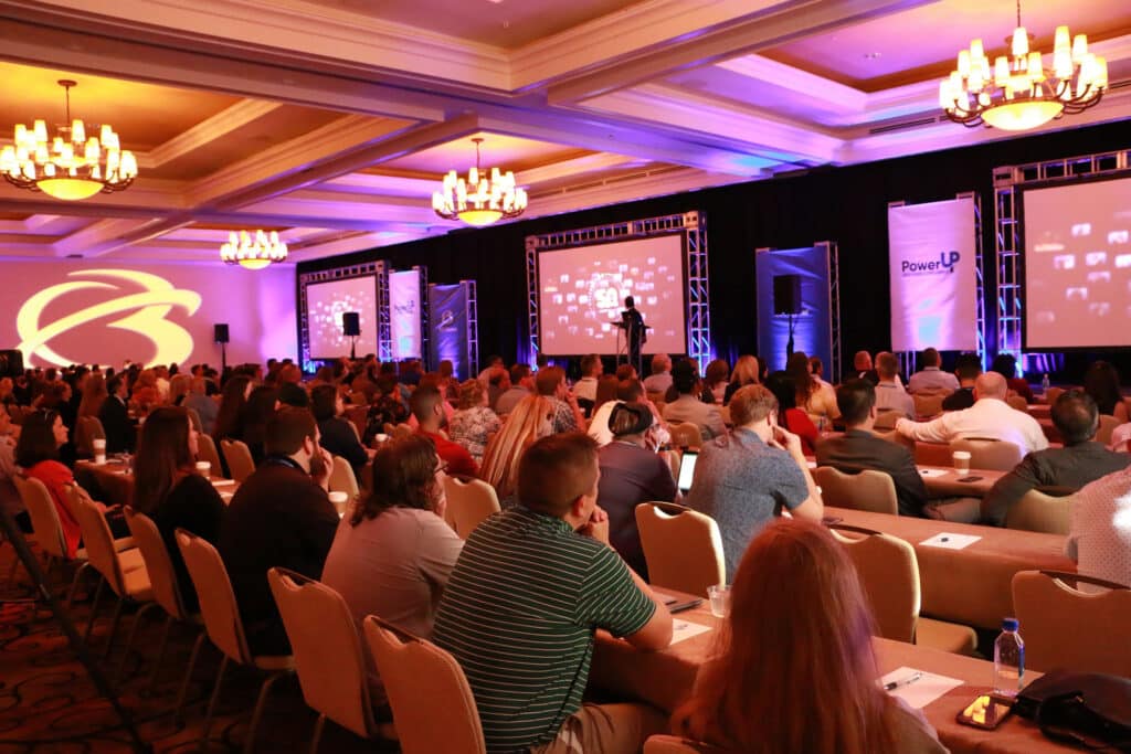 A large audience attends a conference in a ballroom, with a speaker presenting on stage and slides displayed on screens.