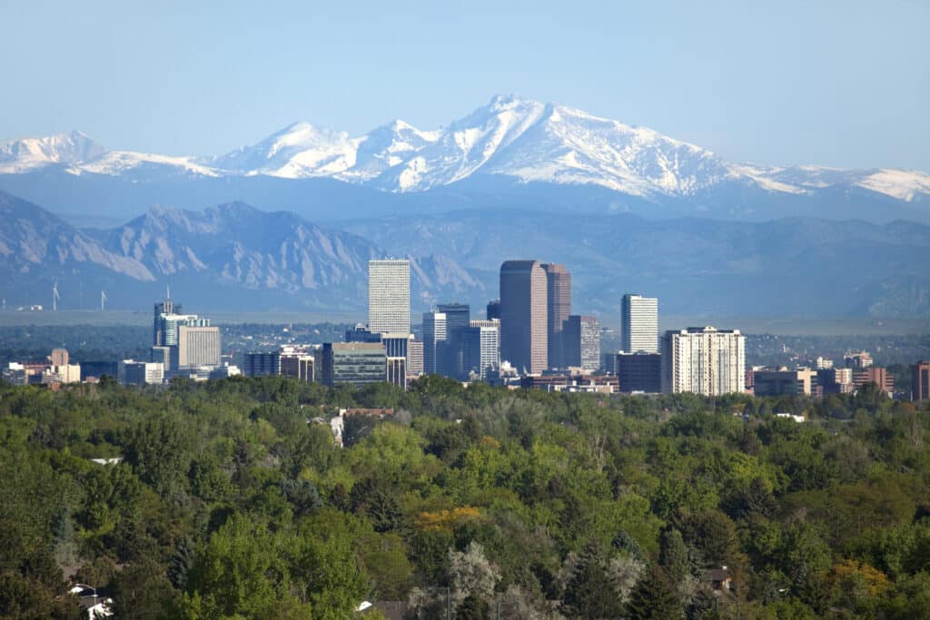 City skyline with tall buildings in the foreground and snow-capped mountains in the background, set against a clear blue sky.
