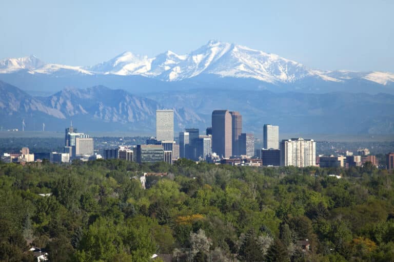 City skyline with tall buildings in the foreground and snow-capped mountains in the background, set against a clear blue sky.