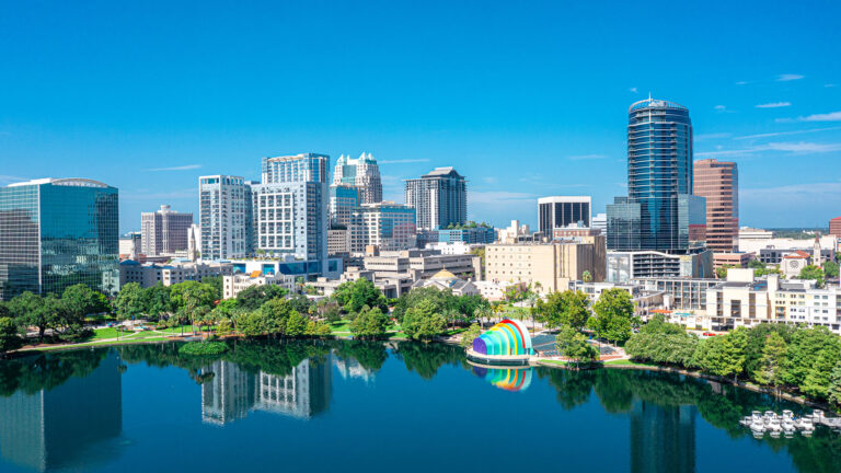 Aerial view of a city skyline with modern skyscrapers, a lake, and a colorful installation near the shore under a clear blue sky.