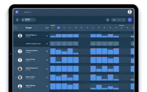 Tablet displaying a scheduling app with a calendar grid, showing people’s names alongside their work hours and shifts highlighted in blue and red blocks for April and May 2023.