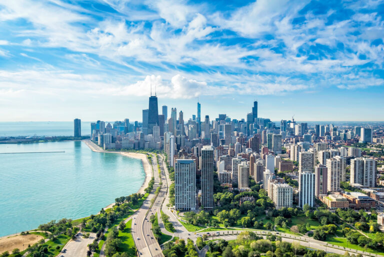 Aerial view of a cityscape with tall buildings, a lakefront, and green spaces under a partly cloudy sky.