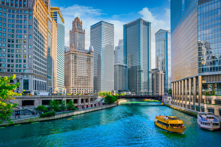 A cityscape with tall buildings flanking a river. A yellow boat sails on the water under a blue sky with clouds.