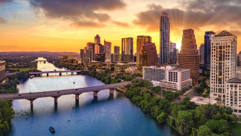Aerial view of a city skyline at sunset with a river and bridge in the foreground, surrounded by green trees and modern skyscrapers.