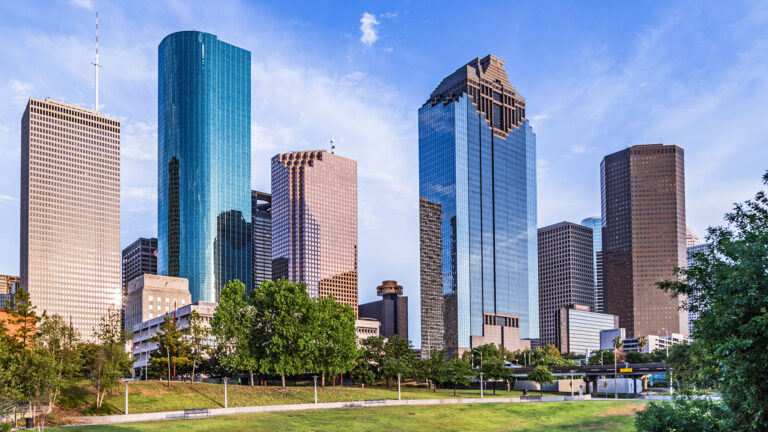 Skyline of a city with modern skyscrapers under a blue sky and trees in the foreground.