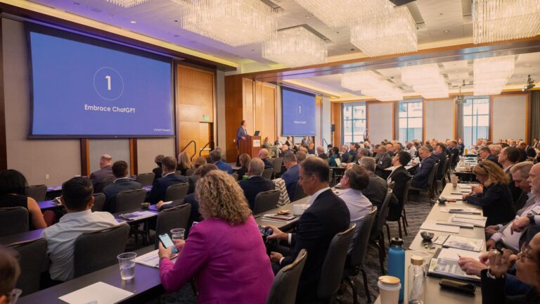 A large audience sits in a conference room watching a presentation with slides titled "Embrace ChatGPT." The room is well-lit with chandeliers.