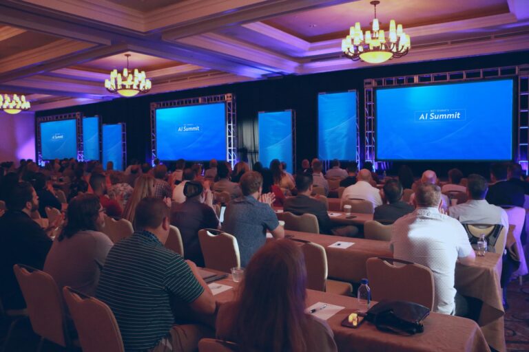 A conference room filled with attendees seated and facing multiple large screens displaying "AI Summit" on stage.