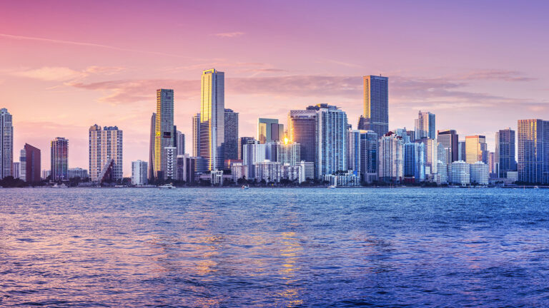 A city skyline with tall buildings at sunset, reflecting in the calm water below.