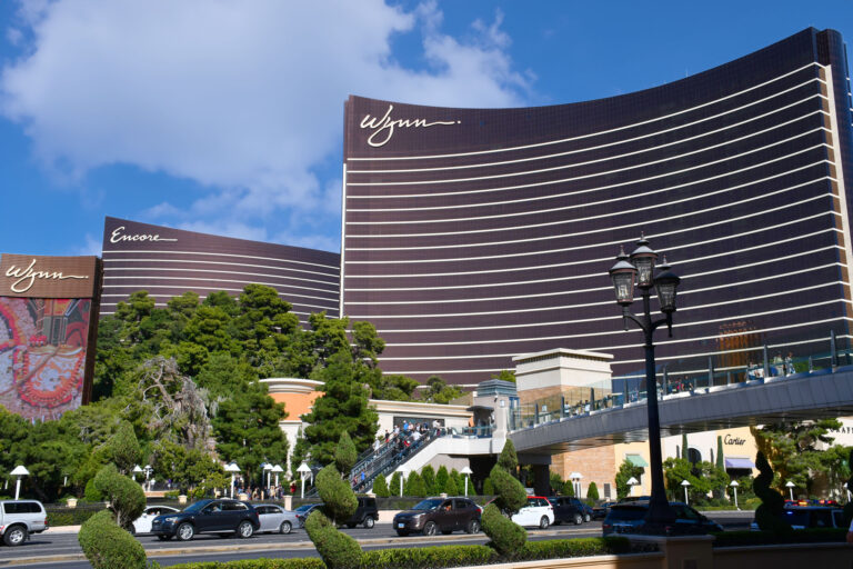 Exterior view of Wynn and Encore Las Vegas hotels with greenery, a pedestrian bridge, and a decorative street lamp in the foreground.