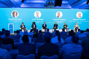 A panel of six people sits on stage with a blue backdrop displaying their names and titles. An audience is seated in front of them.