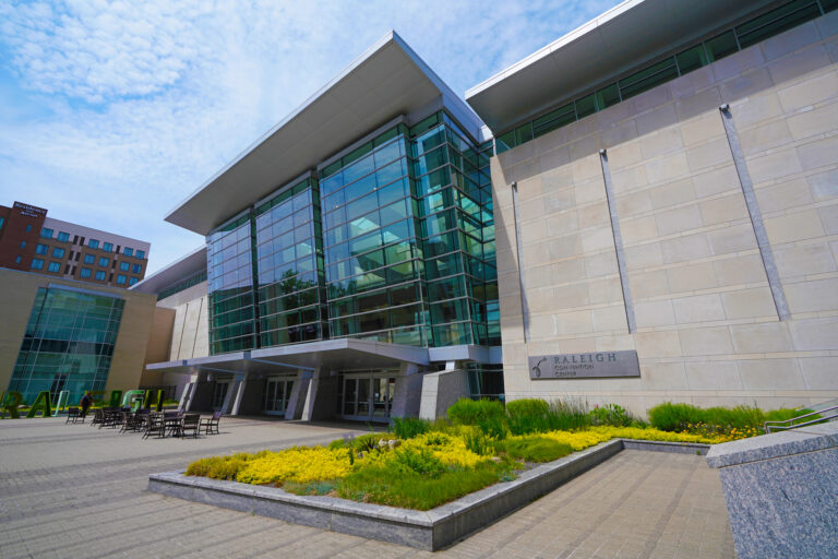Exterior view of Raleigh Convention Center with large glass windows, a stone facade, and a landscaped entrance under a partly cloudy sky.