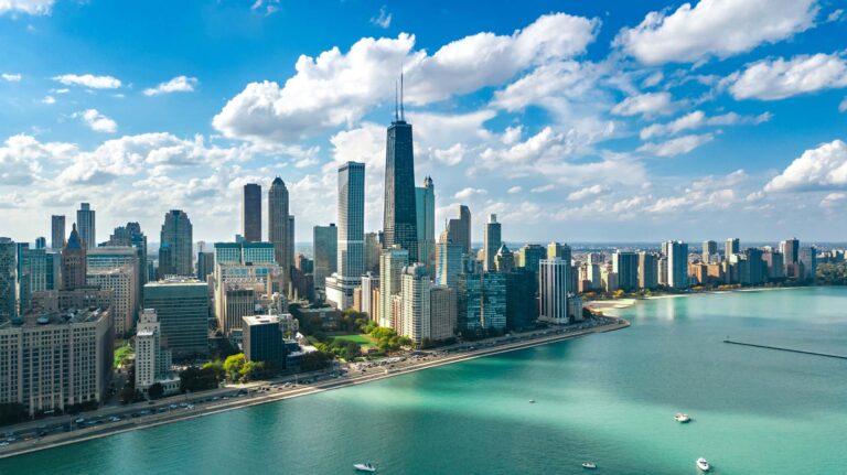 Aerial view of a city skyline with tall skyscrapers beside a large body of water under a partly cloudy sky.