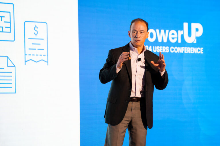 A man in a suit speaks on stage at the PowerUP 2024 Users Conference, with a large screen displaying graphics behind him.