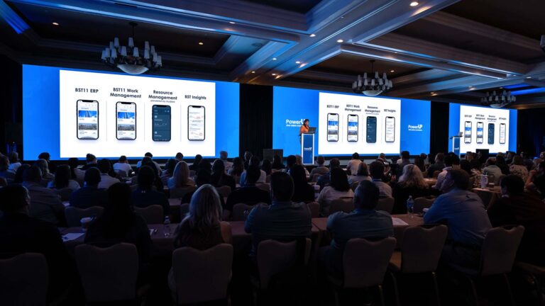 A speaker presenting to a seated audience in a large conference room with three screens displaying app interfaces.