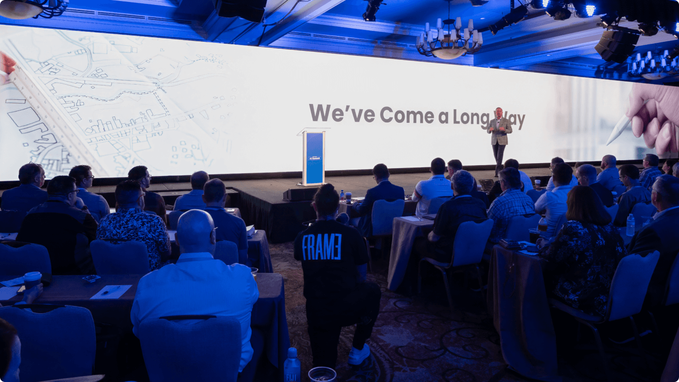 A speaker presents on stage to an audience in a conference room with the words "We've Come a Long Way" displayed on a large screen.