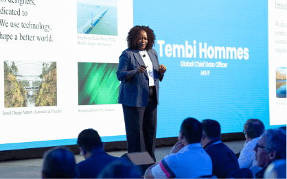 A speaker presents on stage with a large screen displaying slides and the name "Tembi Hommes, Global Chief Data Officer, ARUP." Audience members are seated in the foreground.