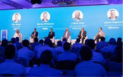 Panel discussion on stage with six individuals seated, five men and one woman, against a blue backdrop showing names and titles. Audience members visible in the foreground.