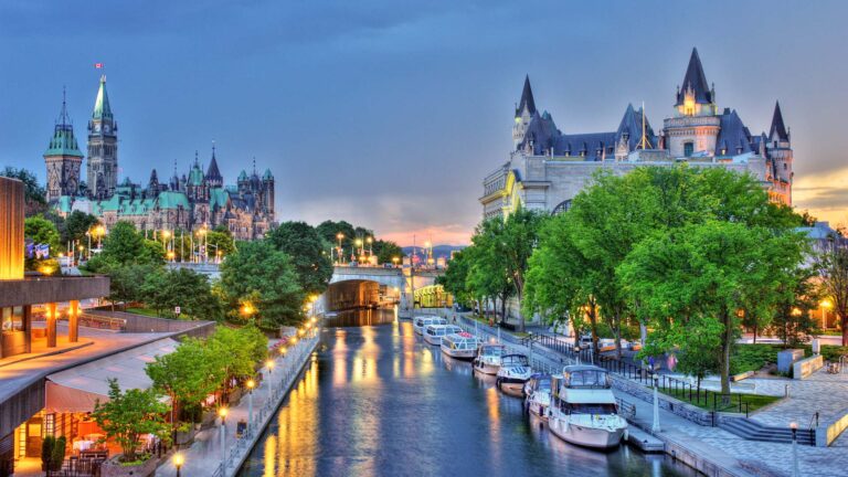 A scenic view of Ottawa featuring the Rideau Canal, flanked by green trees and boats, with historic architecture in the background under a partly cloudy sky at dusk.