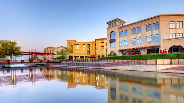 A waterfront view of a modern building complex with outdoor seating, trees, and a bridge, reflected in calm water under a clear sky.