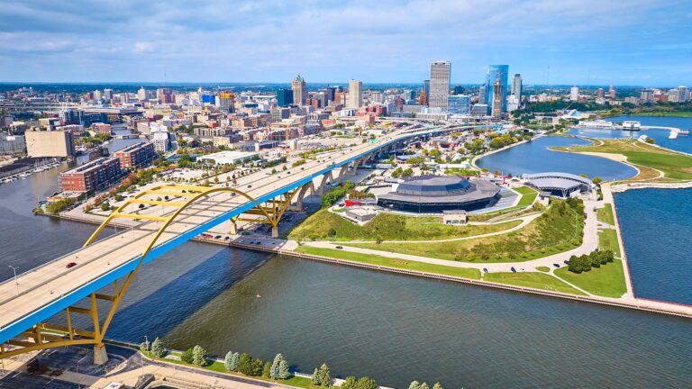Aerial view of a city's waterfront with tall buildings, a curving bridge, and green spaces adjacent to the water.